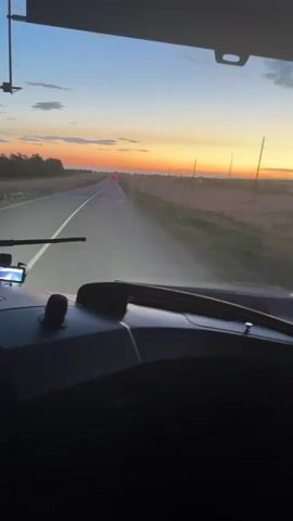 This media file captures a view from the driver's perspective inside a vehicle, likely a truck or bus, as it travels down a road at dusk. The road stretches ahead, leading towards a vibrant sunset painting the sky in hues of orange, yellow, and blue. Silhouetted trees line the left side of the road, while fields and utility poles are visible on the right. The vehicle's headlights are illuminated, cutting through the fading light.

On the dashboard, a GPS device is mounted and visible, suggesting navigation is in use. The interior of the vehicle is dark, with the exception of a screen displaying text that appears to be in Cyrillic. The text translates to "НОЧЬ" which means "night." The time of day is clearly indicated by the sunset. The overall atmosphere is one of travel and transition into the night.

The location context of Бастау, Kazakhstan, suggests a journey through this region of Central Asia. The open, flat landscape with fields and utility poles is consistent with rural or semi-rural Kazakhstan. The emotion conveyed is one of peaceful movement and the beauty of nature as day turns to night.