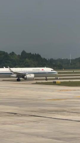 A white Air China passenger airplane with a blue stripe along its fuselage and a red flag on its tail is seen taxiing slowly on an airport runway. The aircraft is moving from left to right in the frame. The word "AIR CHINA" is clearly visible on its side. The runway features concrete pavement with yellow markings, including "C2" and directional arrows. In the background, there are green, forested hills or mountains under a grey, overcast sky. The scene appears to be an airport in Jianyang City, China. No people are visible in the video.