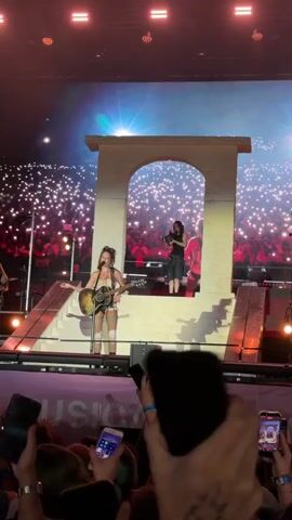 The video shows a live musical performance on a large stage. In the foreground, a crowd of people are holding up their phones, capturing the event. The stage itself features a large screen displaying an image of a performer, likely the one on stage, playing a guitar. There is a white, arched structure center stage, with two performers on elevated platforms behind it. The performer on the left is a woman playing an acoustic guitar and singing into a microphone. She is wearing shorts and knee-high socks. The performer on the right is standing and appears to be playing a violin. The stage is lit with bright lights, and the background screen shows a projection of a stage setup with a giant archway. The overall atmosphere appears to be a concert or festival.