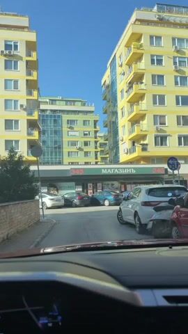 The video, shot from inside a vehicle, captures a street scene in Sofia, Bulgaria, on a bright, sunny morning, indicated by the clear blue sky and the time "8:34" visible on the car's navigation screen. The car's dashboard displays a temperature of "27°C" and air conditioning settings.

The main focus is a commercial building on the ground floor, flanked by two multi-story residential buildings. The building to the right is prominently yellow with green balconies, while the one to the left is light green with blue glass elements.

The commercial storefront features a prominent green facade with the numbers "345" on either side and the Cyrillic word "МАГАЗИНЪ" (Magazin, meaning "store" or "shop") spelled out in large white letters in the center. Another smaller sign on the left side of the storefront reads "DETELINAS".

Several cars are parked in front of the store, including a dark grey sedan, a blue sedan, and a light blue hatchback. A white car is partially visible on the right side of the frame, possibly parked or moving slowly. The vehicle from which the video is recorded has a dashboard in the foreground, showing part of its infotainment screen which displays a map with various Bulgarian street names such as "ул. Света Марина," "ул. Ладога," and "ул. Пирин."