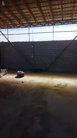 The video shows a mechanic working on the side of a white truck inside a workshop. The mechanic is kneeling and appears to be using tools. The workshop has a concrete floor and a corrugated metal roof. There are several large tires and some buckets visible in the background. The interior of the truck's cab is also shown, revealing the driver's seat, steering wheel, and dashboard.