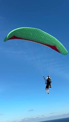 The video shows two people paragliding against a clear blue sky, with a view of the ocean visible in the distance. The main focus is on the paraglider closer to the camera, which has a green and red canopy. The person controlling the paraglider is wearing a white shirt, dark shorts, and white sneakers. Another paraglider, also with a green and red canopy, is seen further away and slightly above the first one. The background is dominated by the bright blue sky, with a small portion of the ocean visible near the horizon.