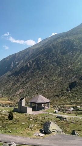 The video shows a mountainous landscape under a clear blue sky with some white clouds. A steep, grassy mountain dominates the background, with rocky outcrops and sparse vegetation. In the mid-ground, there is a small, octagonal covered structure with a dark roof and open sides, supported by pillars. To the left of this structure is a plain, light-colored concrete building with a flat roof and an open section. The ground in the mid-ground and foreground is covered in grass, rocks, and some smaller bushes. A paved road or pathway curves through the scene in the foreground, with some large boulders visible near its edge. In the distance, on the grassy area, there appear to be a few vehicles, possibly campers or vans. The overall scene is one of a natural, rugged environment with man-made structures providing some shelter or facilities.