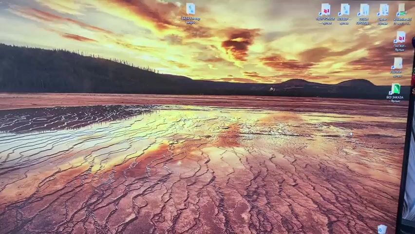 The video captures a computer monitor displaying a landscape image. The image features a vast, colorful hot spring or geothermal pool with intricate, patterned mineral deposits in the foreground, reflecting a dramatic sunset or sunrise sky with vibrant orange, yellow, and red clouds. In the background, a dark, forested hillside and distant mountains are visible. The monitor screen itself shows a typical desktop interface with icons arranged along the top and right edges. There is no discernible action or interaction occurring within the video; it is a static display on a screen.