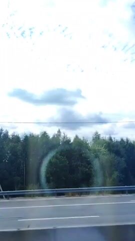 The video captures a view from a moving vehicle on a road. In the foreground, there is a gray concrete barrier and the road itself, with white lane markings visible. A metallic guardrail runs along the edge of the road. Beyond the guardrail, a dense line of green trees forms the backdrop. The sky above is a mixture of bright white and gray clouds, with some patches of blue sky visible. A faint circular light artifact is present in the lower left and center of the frame, suggesting a lens flare or reflection. In one segment, a white box truck is seen driving past on the road. A tall, thin pole with a dark object at the top, possibly a light or sensor, is also visible against the sky. The overall impression is a roadside view on a cloudy day.