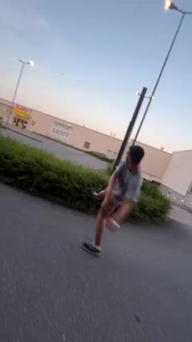 The video captures a brief, dynamic scene outdoors, likely in a commercial or urban fringe area of Oelde, Germany. The time of day appears to be late afternoon or early evening, with the sky showing hues of blue and a hint of sunset. The weather seems clear.

The primary subjects are two young men, though their features are somewhat blurred due to motion. The first individual, in the foreground, is wearing a grey t-shirt and patterned shorts. He is in motion, possibly skateboarding or running, as his legs are spread apart and his body is angled. He glances towards the camera with a neutral expression.

The second individual, further back and to the right, is also in motion and appears to be wearing a grey t-shirt and plaid shorts. Their movement is more blurred, but they seem to be looking in the same general direction as the first person.

The setting includes a paved pathway or road, bordered by a dense, well-maintained green hedge. In the background, there is a large, light-colored building, likely a commercial establishment, with a sign that has some visible text. One sign prominently displays "GETRÄNKE 59302" along with other German text which is partially obscured but includes "IN DER DEZENTRALEN". Another sign, possibly for a store, reads "SEDE". Streetlights are visible, suggesting the area is well-lit or equipped for evening hours.

The activity appears to be casual movement or play, possibly a short video recording of individuals enjoying themselves outdoors. The overall mood is energetic due to the motion, but the facial expressions, though briefly visible, are not strongly indicative of specific emotions. The location cues, such as the building type and the German text, firmly place the scene in Oelde, Germany.