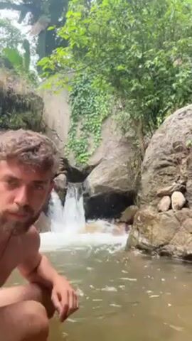 The video shows a shirtless man with a beard kneeling in a shallow, murky body of water with a small waterfall in the background. The waterfall is formed by water cascading over rocks, and the surrounding area is lush with greenery, including trees and vines. There are large boulders on either side of the waterfall. The man is looking directly at the camera at the beginning, then turns and submerges himself further into the water.