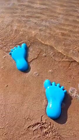 This is a close-up video of two bright blue, cartoonish foot-shaped objects resting on a sandy beach. The objects are submerged partially in shallow, clear water that is lapping at the shore. The water is creating small waves and ripples around the foot shapes, and some tiny bubbles are visible. The sand is a light brown color and appears damp where the water has touched it. The lighting suggests it is daytime, possibly a sunny day, casting subtle shadows. The objects are positioned at an angle to each other, with one slightly further up the beach than the other.