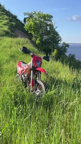 A red dirt bike is initially parked in tall green grass on a hillside. The camera pans right, revealing a wide body of blue water below the hillside. A long bridge with multiple piers spans the water in the distant background. The hillside is covered in green grass and some trees, with visible eroded slopes leading down to the water's edge. The sky above is blue with scattered white clouds. The water surface has small ripples. No individuals are visible.