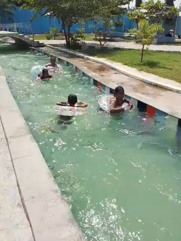 The video shows four people floating in a narrow, shallow canal. The water in the canal is a light greenish-blue. Each person is using a colorful inflatable ring to stay afloat. The canal has concrete edges, and there is grass and trees visible along the sides. In the background, there are blue buildings and a small bridge. The people in the canal appear to be enjoying themselves.