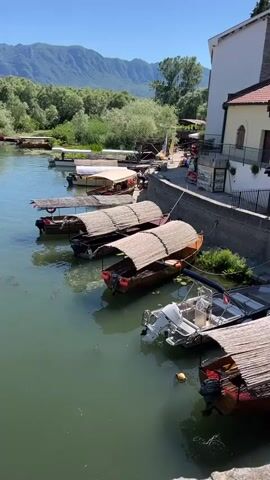 The video pans across a body of water, likely a river or lake, lined with several boats docked along a stone embankment. The boats are of varying sizes and types. Some are small, traditional wooden boats with thatched roofs, while others are larger, motor-powered vessels. The water appears green and slightly murky, with some aquatic vegetation visible on the surface.

The embankment itself is made of rough stone and has a railing along the top. Beyond the embankment, a path or road runs parallel to the water, with some small buildings and structures, including what appears to be a small cafe or shop. Trees and lush green vegetation line the opposite bank of the water, extending up a gentle slope towards distant mountains that are shrouded in a light haze. The sky is clear and blue, indicating a sunny day.

There are a few people visible in the video, though their actions are not prominent. They appear to be casually moving along the embankment or near the docked boats. One person is seen in one of the larger motorboats, possibly preparing for departure or having just arrived. The overall atmosphere is peaceful and suggests a tourist or recreational area. The camera movement is smooth, providing a sweeping view of the entire scene.