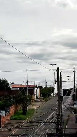 The video begins with a view of a street bordered by small buildings under a cloudy sky, with a plane flying overhead. The camera then pans to the right, revealing a rooftop where clothes are hanging on a clothesline. A woman with long dark hair, wearing a green shirt and patterned leggings, walks into the frame and past the clothesline.