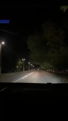 A first-person perspective video shows a car driving down a road at night. The road is lined with streetlights and trees. The headlights of the car illuminate the road ahead. There are a few other cars visible in the distance. The video appears to be taken from inside the car, looking out the windshield.