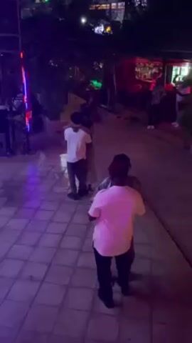 In an outdoor space at night, with a bar visible in the background, four people are dancing in pairs. The lighting is purple. The pair on the left consists of two people wearing white shirts, one in dark pants and the other in dark grey pants. They are holding each other closely and swaying to the music. The other pair consists of a person in black pants and a pink top, and another in a patterned shirt and dark pants, holding each other and swaying. The floor is tiled. There are trees and a vertical structure with red and blue lights in the background. Other people are standing and chatting near the bar.