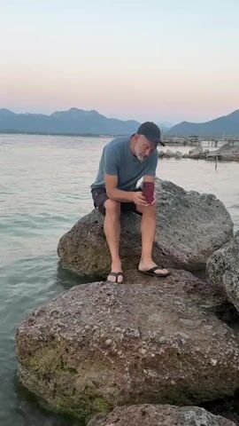 A man in a blue t-shirt and shorts sits on large rocks by the water, looking down at his phone. In the background, there are mountains and a few people in the water. The sky is a soft pink and purple.