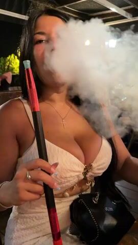 The video shows a woman with dark hair and a white dress smoking a hookah. She is holding the hookah hose and exhaling smoke. In the background, there are several other people sitting at tables in what appears to be an outdoor restaurant or bar. There are drinks on the tables, including what looks like a mojito and a darker colored drink. One man in the background is looking at his phone, and another man is making a peace sign. The overall atmosphere appears to be casual and social.