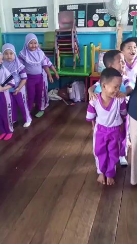 A group of young children, dressed in matching purple and white uniforms, are standing in a line, facing the camera. The children are holding blue balloons between their knees. The room appears to be a classroom, with colorful decorations on the walls, shelves filled with books and toys, and wooden floors. The children seem to be participating in a game or activity. The overall atmosphere is cheerful and playful.