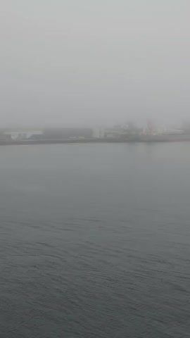 This is a still frame from a video taken from a ship at sea. The view is of a hazy, gray expanse of water, with a faint, out-of-focus industrial landscape visible on the horizon in the upper left corner. The water is calm with gentle ripples. The overall atmosphere is one of limited visibility due to fog or mist.