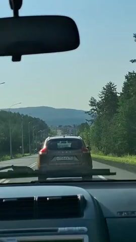 The video shows a view from inside a car traveling on a road. The camera is positioned behind the dashboard, facing forward through the windshield. A rear-view mirror is visible at the top of the frame. In front, another car, a brown hatchback, is driving on the same road. The road is bordered by trees and what appear to be streetlights on the left side. In the distance, rolling hills or mountains can be seen under a clear sky. The overall impression is a journey through a scenic, possibly rural or suburban, landscape.