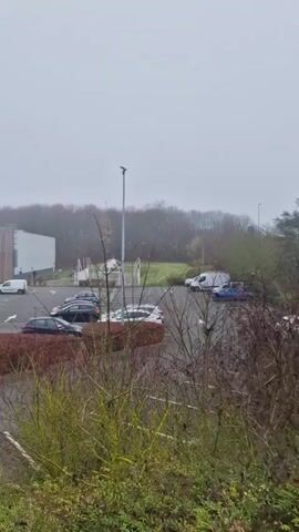The video shows the outside of a Decathlon store on a cloudy day. The store is a large, rectangular building with a light brown wooden façade and the Decathlon logo prominently displayed. In front of the store is a parking lot with several cars parked in it. A few people can be seen entering and exiting the store. There is a patch of greenery in the foreground. The camera then pans to the left, showing a road with a crosswalk. A few cars are driving on the road. A metal guardrail runs along the side of the road, with some vegetation growing between it and the road.