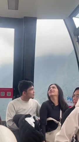The video shows the interior of a cable car with several people inside. In the foreground, a young man and woman are seated next to each other, looking up and to the right. The man is wearing a cream-colored sweatshirt and the woman has long dark hair and is wearing a black top. To their right, partially visible, is another person. In the background, through the large window of the cable car, mountains can be seen shrouded in fog or clouds. Further to the left, other passengers are seated, including individuals wearing brown robes and yellow caps with a red symbol. The cable car appears to be in motion, ascending or descending a mountain. A sign with the number 31 is visible on the window.