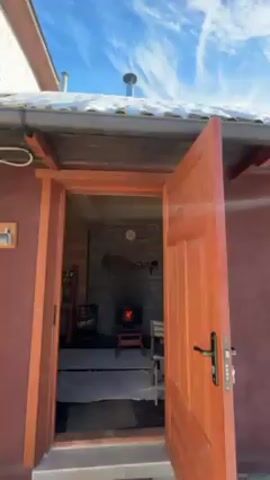 The video begins looking into a room through an open door. The room contains a wood burning stove, a clock, some wooden furniture, and a white rug. The ceiling is made of wood and the back wall of the room is made of grey stone. The video then pivots upward, focusing on the roof of the building and a metal chimney. The roof is covered in snow and the sky is blue with light clouds.