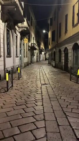 The video shows a nighttime view of a narrow cobblestone street lined with buildings. A streetlamp illuminates the scene, casting shadows on the ground. There are no people or vehicles visible in the frame.