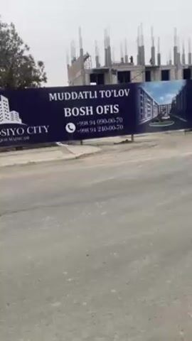 The camera moves to show a street with a large blue sign for Gala Osiyo City, displaying contact phone numbers and a photo of the planned buildings. Behind the sign, construction is underway, and figures that may be workers can be seen at the top. The sky is overcast, and there are trees in the background. The street has some cracks and imperfections.
