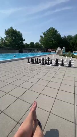 A low-angle, first-person perspective shot shows a person's bare legs and feet resting on a tiled patio. In the background, a large outdoor chess set is arranged on the patio. Beyond the chess set is a swimming pool with several people swimming and relaxing. Trees and a bright blue sky with some clouds form the backdrop.