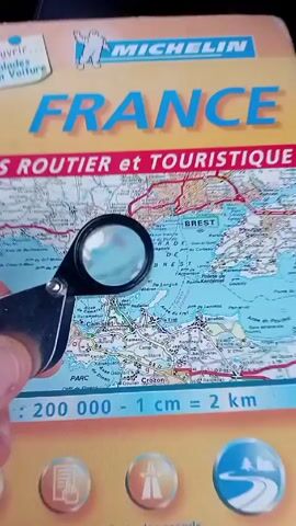 Here is a concise description of the video:

The video shows a map of France.  A hand holds a magnifying glass over different parts of the map, allowing closer inspection of the smaller towns and details, for example, zooming in on Kernao. The magnifying glass has a black frame.  The map is colorful, with terrain depicted in green, blue, and white. There are red lines indicating roads. Text on the map is in French.