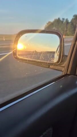 The video shows a view from inside a car as it drives along a road during sunset. The sun is low in the sky, casting a golden light and creating lens flares. The side mirror reflects the sunset and the road ahead. Trees and a field are visible in the background. The interior of the car, including the door panel and window, is also visible. The video is short and shows a continuous movement.