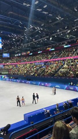 The video shows a packed indoor arena with figure skaters on the ice. The crowd fills the stadium seats, surrounding the rink marked with "Milano Cortina 2026" and the Olympic rings. Two pairs of figure skaters stand on the ice, one pair in black and blue outfits and another pair in green and black. They appear to be acknowledging each other or coaches. The overhead lighting is bright, with a large screen displaying video above the rink. Several photographers stand close to the ice. The camera is held by an audience member, providing a slightly obstructed view from their seat.