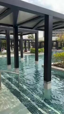 The video shows a swimming pool area at a resort. The camera is positioned under a shaded structure with black pillars that extend into the water. The pool is large, tiled, and surrounded by landscaping including small islands of green plants and flowers. In the background, there are lounge chairs with white umbrellas and other shaded structures around the pool. People are swimming and lounging in the pool. The sky is overcast.
