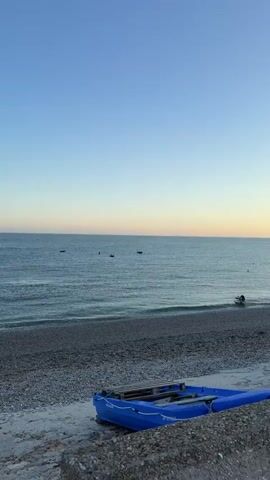 A blue inflatable raft rests on a pebbled beach, partially submerged in the gentle waves of the ocean. In the distance, a small white motorboat is positioned near the shore, with a person leaning over its stern. The sky is a clear, soft gradient of blue fading into a pale orange hue near the horizon, indicating either sunrise or sunset. Several buoys are visible in the water, and distant, silhouetted wind turbines dot the horizon.