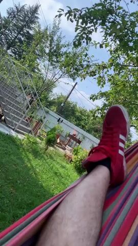 The video shows a person's legs and feet wearing red sneakers with white stripes, resting in a hammock. The background is a green yard with trees and a fence. There is a barbecue grill in the background with fire. The sky is visible and appears to be blue with some clouds. The perspective is from someone lying in the hammock, looking out at the yard.