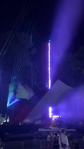 The video shows an outdoor event at night. A stage is set up with a large, abstract geometric backdrop featuring red and white panels. Various lights are illuminating the stage and the surrounding area, including blue and purple lasers that create beams of light in the smoky atmosphere. On the stage, there are speakers and what appears to be a DJ booth with at least two people operating equipment. In front of the stage, a crowd is gathered, separated from the performance area by a metal fence. Several individuals are visible in the foreground, some facing the stage and others turned away from the camera. The overall scene suggests a music festival or concert.