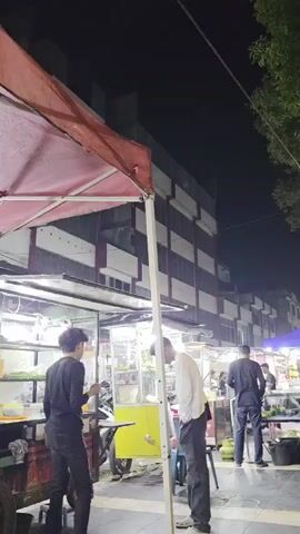 The video shows a nighttime scene of a street food vendor area. Several food stalls are visible, set up under awnings and tents. People are milling about, some appearing to be customers and others working at the stalls. One person in a white shirt and black pants stands facing a stall, looking to their left. Another person in a black shirt and dark pants is seen from behind, also facing a stall. Further down the street, multiple other stalls are illuminated by lights, with people preparing food and interacting. The background shows a multi-story building with dark windows and balconies. Trees with dense foliage line the right side of the frame. The ground appears to be paved. A yellow cart is parked near the foreground stall. Gas tanks are visible near some of the stalls. A person wearing a hijab sits on a chair at one of the stalls.