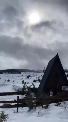 The video showcases a winter landscape featuring a triangular-shaped wooden cabin in the foreground. The cabin, with its dark roof and wooden facade, is situated in a snow-covered area dotted with small trees and what appears to be fencing. In the background, a mountain range is visible, partially obscured by a blanket of grey clouds. The sun attempts to pierce through the cloud cover, casting a soft, diffused light over the scene. There are power lines visible in the distance. No people are visible.