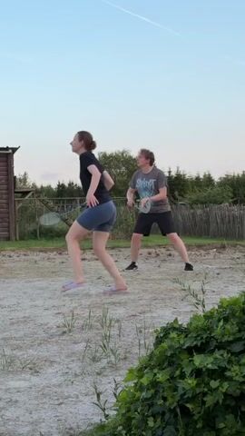Two individuals are playing badminton in an outdoor area. The person in the foreground, identified as a young female, is wearing a dark t-shirt and blue shorts, with light-colored flip-flops. She is in motion, reaching to hit a badminton shuttlecock with a racket. Behind her, a second individual, likely a young male, is also playing badminton, wearing a grey t-shirt and dark shorts, with dark sneakers. He is also in motion, having just hit the shuttlecock. The environment appears to be a dusty or sandy court surrounded by grass and bushes. In the background, there is a wooden structure and a fence, with trees and a clear sky with a contrail overhead. The lighting suggests it is late afternoon or early evening.