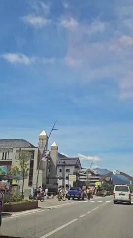 A white van drives down a street in a town. There are buildings on both sides of the street. The sky is blue with some clouds. There are people walking and riding bicycles on the sidewalk. There is a bridge or archway over the street.