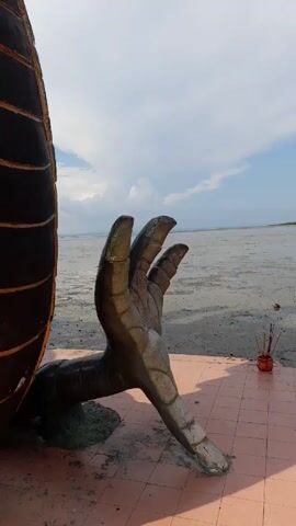The video shows a statue of a hand, possibly made of concrete or stone, with its fingers pointing upwards. The hand is weathered and has a dark, aged appearance. To the right of the hand, there is a red flower pot containing incense sticks. The foreground is a tiled surface, and in the background, there is a muddy expanse that stretches to the horizon under a sky with scattered clouds. The environment appears to be a coastal or waterfront area with visible tidal flats.