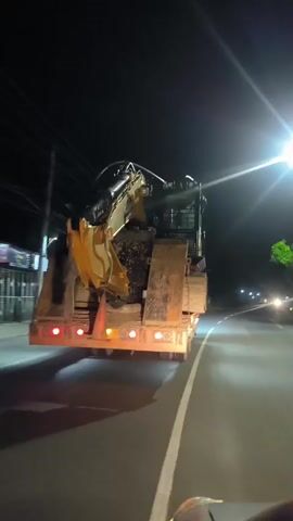 The video shows a yellow excavator being transported on a flatbed trailer at night. The excavator is loaded onto the trailer, facing backwards. The trailer has red brake lights and orange turn signals. The excavator appears to be covered in dirt. The trailer is on a road with streetlights and power lines visible in the background. There are also some trees and buildings on the side of the road. The video is shot from a moving vehicle behind the trailer.