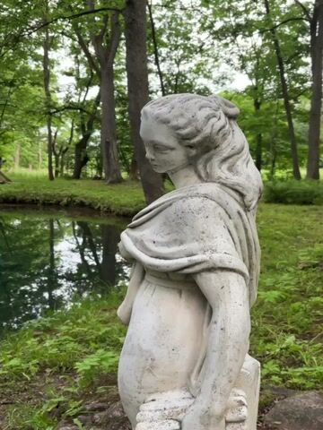 The video shows a close-up view of a weathered stone statue of a woman in a garden setting. The statue is positioned in the foreground, facing left, with its back to the viewer. The woman is depicted with her hair pulled back and draped in flowing robes. Her expression is pensive, and her gaze is directed downward. In the background, a serene pond reflects the surrounding lush green trees and foliage. A wooden structure, possibly a small bridge or viewing platform, is partially visible on the left edge of the pond. The overall atmosphere is tranquil and somewhat melancholic, suggesting a secluded natural space with a touch of classical art. The lighting is soft and diffused, indicating an overcast or shaded environment.