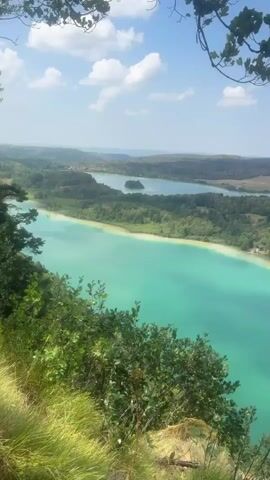 A high-angle shot captures a vibrant turquoise lake surrounded by lush green trees and rolling hills under a partly cloudy blue sky. The camera pans slowly, revealing the expanse of the lake and the verdant landscape. In the foreground, the camera is partially framed by green leaves and branches. The water of the lake exhibits a striking color contrast, with clear turquoise in some areas and a lighter, sandy hue closer to the shore. The surrounding terrain features a mix of dense forests and open, grassy fields. The scene is tranquil and natural, with no signs of human activity or structures immediately visible in the shot.