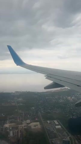 The video shows a view from an airplane window. The plane's wing is visible, with a blue-painted tip. Below, a city extends along the coastline. The body of water is partially obscured by haze. Overcast skies dominate the upper portion of the frame.