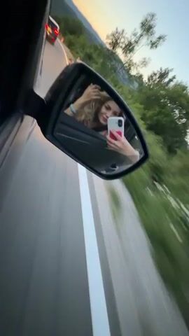 The video shows a view of a road from the side mirror of a car. The car is moving quickly, as evidenced by the blurred lines on the road and the moving scenery. In the side mirror's reflection, a woman with wavy, blonde hair is visible. She's holding a phone, possibly taking a selfie or recording a video, and adjusting her hair. The background in the mirror's reflection includes trees, mountains, and a twilight sky. Another car is visible ahead on the road. The overall impression is of someone enjoying a road trip, capturing a moment on camera.