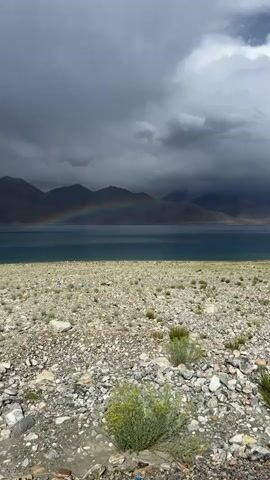 The video captures a panoramic view of a vast, rocky terrain leading to a serene lake. In the distance, majestic mountains loom under a dramatic sky filled with dark, ominous clouds. A vibrant rainbow arches gracefully across the sky, bridging the gap between the mountains and the water, adding a touch of surreal beauty to the otherwise rugged landscape. The lake's surface reflects the colors of the sky, creating a striking contrast with the muted tones of the land. Small green shrubs dot the rocky ground, adding hints of life to the otherwise desolate scene.