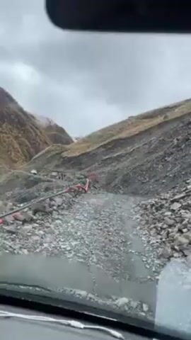 The view from inside a vehicle shows a rocky, mountainous environment on an overcast day.  The ground is covered in loose rocks and gravel, with some water visible on the surface. Red industrial hoses are laying on the ground. The terrain is steep and uneven. A small white vehicle can be seen farther up the road.  The windshield shows water droplets on the surface obscuring the image.