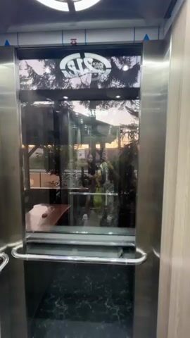 The video shows an interior view of a glass elevator cabin with stainless steel walls. The elevator is moving upwards, and the camera is positioned at the front of the cabin, looking through the glass doors. Reflections on the glass doors show a sunset with trees and buildings. A person is visible in the reflection, appearing to take the video. The elevator cabin has a chrome handrail and a black and white patterned floor. Above the glass doors, there is a logo that appears to read "ATS 1994 HOPA".