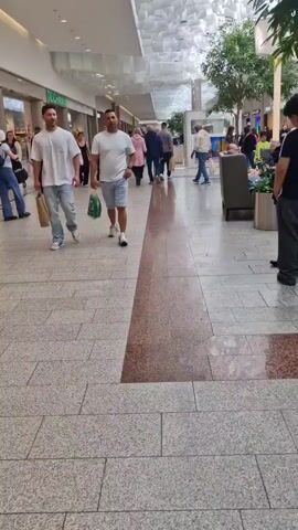 The video is filmed from a low angle, looking down a shopping mall corridor. The floor is tiled with large grey tiles and a central strip of speckled brown and red tiles. The ceiling is made of a white, textured material that appears to be a grid of abstract shapes. People are walking in both directions. In the foreground on the left, two men in white t-shirts are walking away from the camera. The man on the right is carrying a green and white shopping bag. To the right of the central tile strip, a man in a white t-shirt and blue shorts is walking towards the camera, also carrying a green and white shopping bag. Further down the mall, other shoppers are visible, including a woman pushing a stroller, a child in a bright yellow jacket, and people sitting on a bench to the right. Trees and plants are visible in planters along the right side of the corridor. The lighting is bright and appears to be artificial, suggesting an indoor environment.